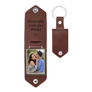 May include: A brown leather keychain featuring a metal snap closure and a square photo insert showing a couple. The interior is engraved with the text Drive safe, Love you always and a heart symbol. A matching brown leather key fob is included.