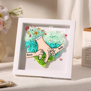 May include: A white framed artwork featuring a heart-shaped arrangement of teal and green flowers. The artwork includes illustrations of hands holding each other, with the names "Annie" and "Bella" on the hands. The text "Mummy WE LOVE YOU" is also present.