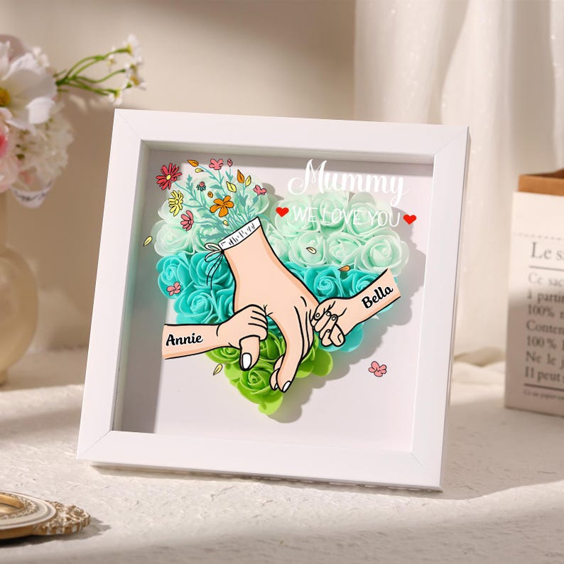May include: A white framed artwork featuring a heart-shaped arrangement of teal and green flowers. The artwork includes illustrations of hands holding each other, with the names "Annie" and "Bella" on the hands. The text "Mummy WE LOVE YOU" is also present.
