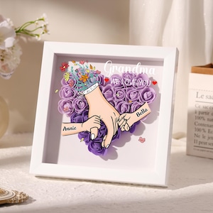 May include: A white framed artwork featuring a heart-shaped arrangement of purple paper roses. The artwork includes illustrations of hands holding each other, with the names "Annie" and "Bella" on the hands. The text "Grandma WE LOVE YOU" is also displayed.