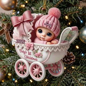 May include: A Christmas tree ornament shaped like a white and pink baby carriage featuring a pink bow, a small pink rose, and the text 2026 and Baby's 1st Christmas. Inside sits a baby wearing a pink knit hat with the name Lucy embroidered on it.