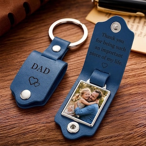 May include: A blue leather keychain with a metal ring and snap closure. The exterior is engraved with DAD and two hearts. The interior features a square metal frame containing a photograph of a child hugging an adult, with the text Thank you for being such an important piece of my life and a heart symbol.