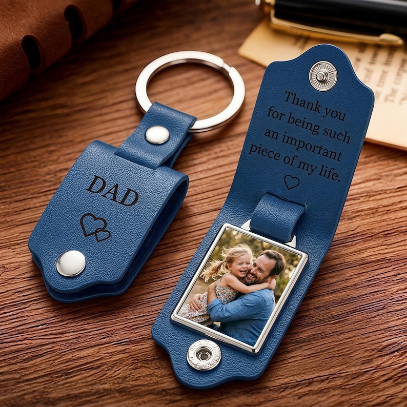 May include: A blue leather keychain with a metal ring and snap closure. The exterior is engraved with DAD and two hearts. The interior features a square metal frame containing a photograph of a child hugging an adult, with the text Thank you for being such an important piece of my life and a heart symbol.