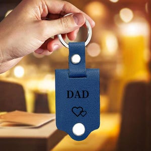 May include: A blue rectangular leather keychain featuring a silver metal ring at the top. The keychain is embossed with the word DAD in black capital letters above two small black heart outlines. A silver metal rivet secures the leather strap.