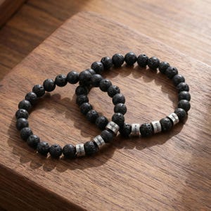 May include: Two black lava stone bead bracelets with silver name beads. The bracelets are on a wooden surface. Each bracelet has a series of black beads and several silver beads with names engraved on them.