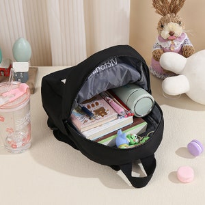 May include: A black backpack with a zipper closure, containing a notebook with a brown bear illustration, a light green insulated bottle, a pink pen, and a small green character keychain. The interior lining is grey, and the word Victory is visible.