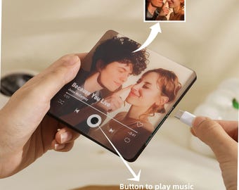 May include: A clear, rectangular music player with a photo of a couple, the song title "Because You Live" and a play button. A small photo of the couple is in the upper right corner. A white USB-C cable is plugged into the device.
