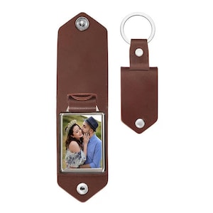 May include: A brown leather keychain featuring a rectangular frame with a photograph of a couple. The keychain has a snap button closure and a silver metal ring. The photograph shows a person with long hair and a floral crown embracing a person in a blue shirt.