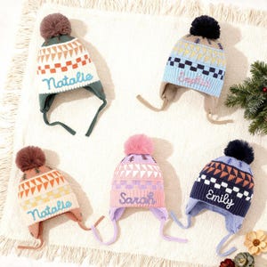 Puede incluir: Cinco coloridos gorros de invierno de punto con orejeras y pompones. Cada gorro presenta una combinación de colores diferente, incluyendo blanco, azul, rosa y marino, con patrones geométricos y los nombres "Natalie", "Sophia", "Sarah" y "Emily" bordados.