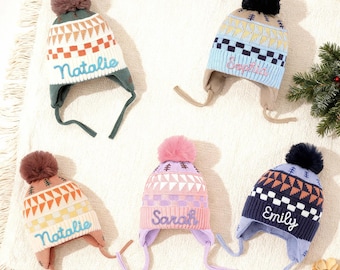 Puede incluir: Cinco coloridos gorros de invierno de punto con orejeras y pompones. Cada gorro presenta una combinación de colores diferente, incluyendo blanco, azul, rosa y marino, con patrones geométricos y los nombres "Natalie", "Sophia", "Sarah" y "Emily" bordados.