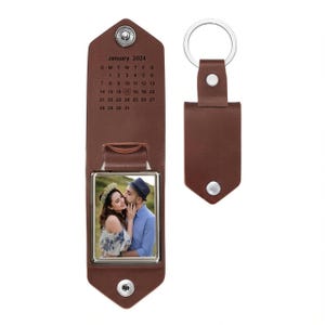 May include: A brown leather keychain featuring a fold-out design with a silver metal snap closure. The interior displays a January 2024 calendar with a heart icon on the 17th and a square frame containing a photograph of two people embracing.