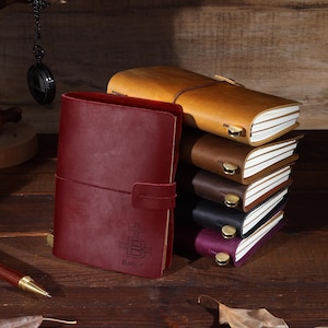 Puede incluir: Una pila de seis diarios encuadernados en cuero con un diario rojo en la parte superior. El diario rojo tiene una cruz y la palabra "Babur" grabada en la portada.