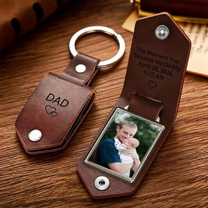 May include: A brown leather keychain with a metal ring, featuring a folded design secured by a silver snap. The exterior is engraved with DAD and two hearts. The interior reveals a photo of a person holding a baby and text: The Moment You Became My Daddy April 20, 2026, 6:25 AM.