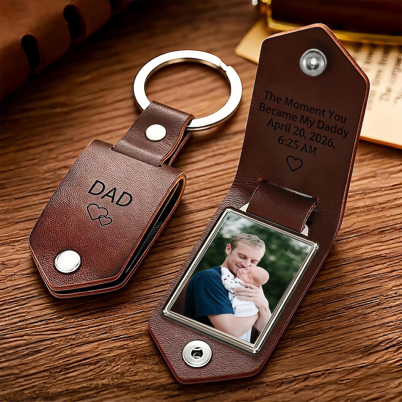 May include: A brown leather keychain with a metal ring, featuring a folded design secured by a silver snap. The exterior is engraved with DAD and two hearts. The interior reveals a photo of a person holding a baby and text: The Moment You Became My Daddy April 20, 2026, 6:25 AM.