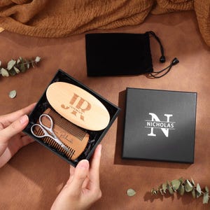 May include: A personalized beard grooming kit in a black gift box. The kit includes a wooden brush with initials and name, a wooden comb, and small scissors. A black drawstring bag and a box with the name "Nicholas" are also included.