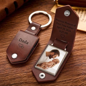 May include: A brown leather keychain with a metal ring, featuring a folded cover secured by a silver snap. The exterior is engraved with "Dada" and two hearts. Inside, a photo of a parent holding a baby is visible, with text reading "Precious Moment of First Time Father April 20, 2026, 6:25 AM" and a heart icon.