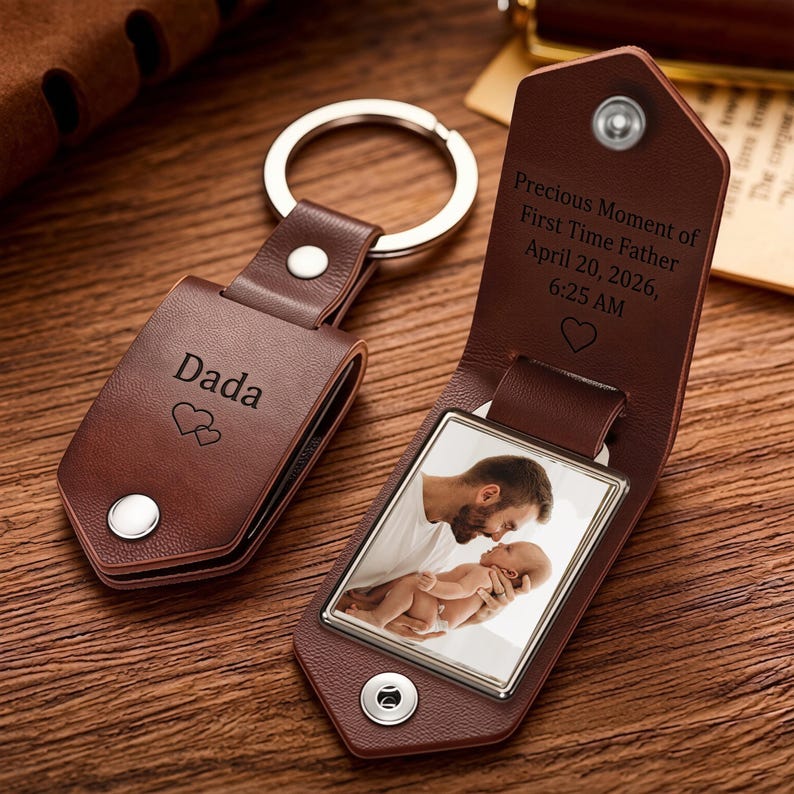 May include: A brown leather keychain with a metal ring, featuring a folded cover secured by a silver snap. The exterior is engraved with "Dada" and two hearts. Inside, a photo of a parent holding a baby is visible, with text reading "Precious Moment of First Time Father April 20, 2026, 6:25 AM" and a heart icon.