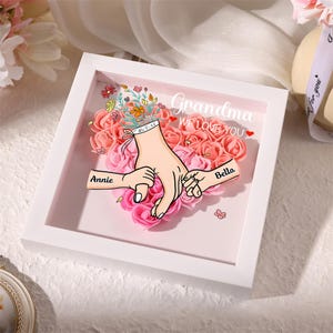 May include: A white framed artwork with a floral heart design. The artwork features a graphic of hands holding each other, with the names "Annie" and "Bella" on the hands. The words "Grandma WE LOVE YOU" are written above the hands.