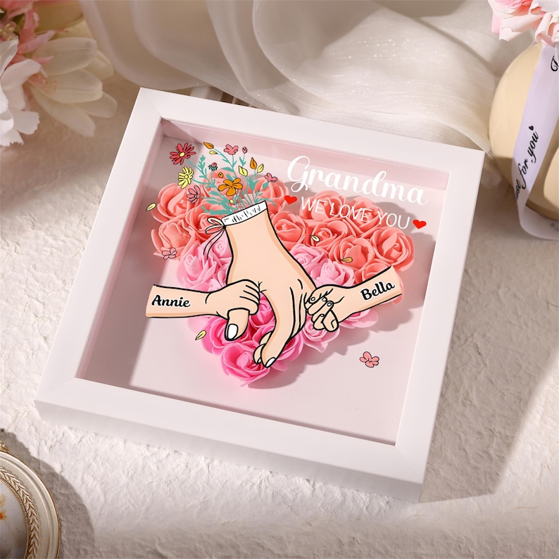 May include: A white framed artwork with a floral heart design. The artwork features a graphic of hands holding each other, with the names "Annie" and "Bella" on the hands. The words "Grandma WE LOVE YOU" are written above the hands.