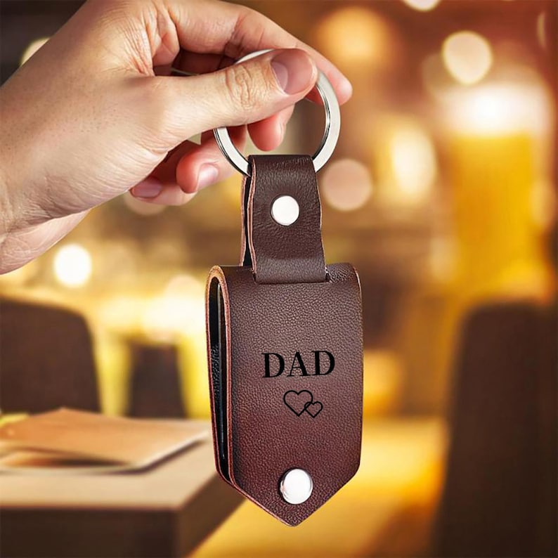 May include: A dark brown leather keychain featuring a silver metal ring and two silver snap buttons. The leather surface is embossed with the word DAD in capital letters above two small heart outlines, creating a simple and functional accessory.