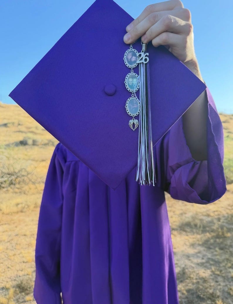 K&ouml;nnte beinhalten: Ein lila Abschlusskappen- und Talar-Set mit einer Gedenkquaste, die einen silberfarbenen Anh&auml;nger mit der Zahl 26, drei ovale Fotorahmen mit jeweils einem kleinen Portr&auml;t und einen silbernen Engelsfl&uuml;gel-Anh&auml;nger aufweist.