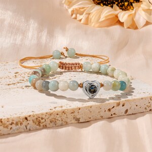 Puede incluir: Un brazalete de cuentas de color verde claro y marr&oacute;n con un dije de coraz&oacute;n plateado. El dije tiene una peque&ntilde;a piedra transparente en el centro.