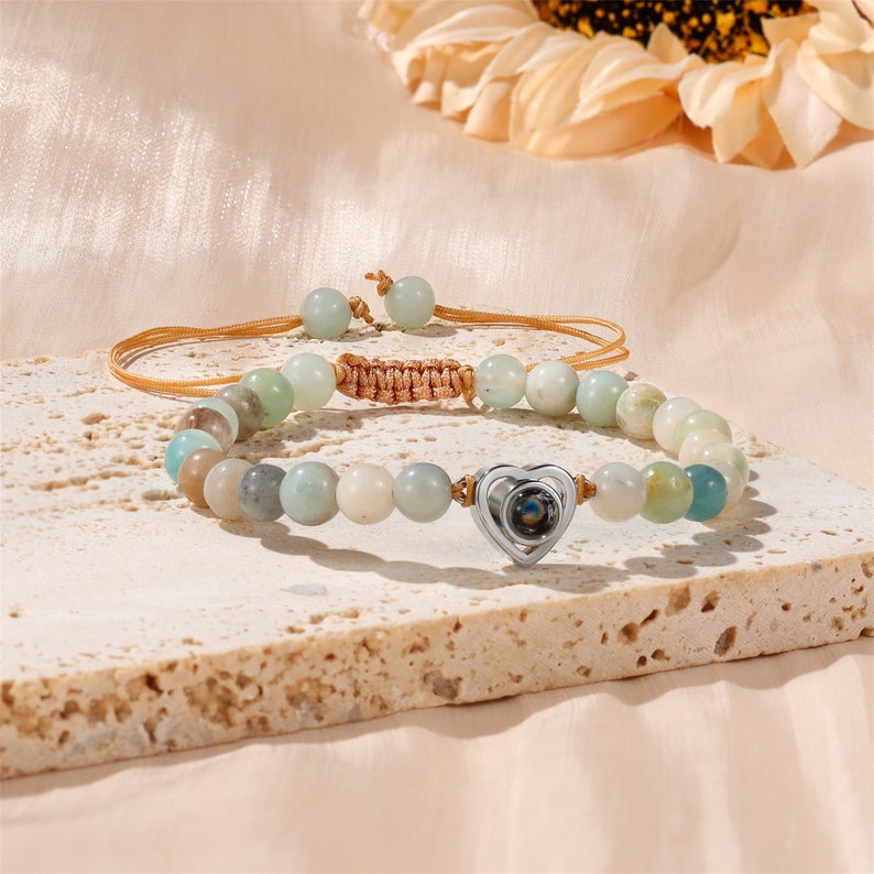 Puede incluir: Un brazalete de cuentas de color verde claro y marr&oacute;n con un dije de coraz&oacute;n plateado. El dije tiene una peque&ntilde;a piedra transparente en el centro.