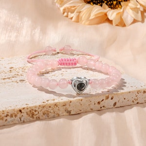 Puede incluir: Un brazalete de cuentas rosas con un dije de coraz&oacute;n plateado. El dije de coraz&oacute;n tiene una peque&ntilde;a piedra negra redonda en el centro.