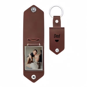 May include: A brown leather keychain with a silver metal ring and a snap closure. The keychain features the word Dad and a heart symbol engraved on the front. Beside it is an open brown leather photo holder displaying a picture of a person holding a baby.