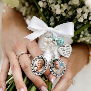 Può includere: Un bouquet da sposa con fiori bianchi e un nastro bianco. Due ciondoli fotografici argentati con accenti di perle pendono dal nastro. Un ciondolo ha un cuore che dice "you are always in my heart."