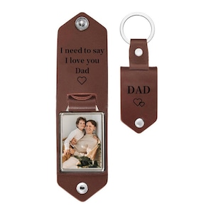 May include: A brown leather keychain featuring a rectangular photo frame insert showing a person holding a child. The leather flap is engraved with the text I need to say I love you Dad and a heart. A separate matching leather tag is engraved with DAD.