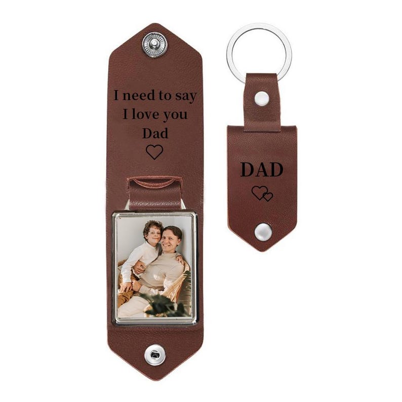May include: A brown leather keychain featuring a rectangular photo frame insert showing a person holding a child. The leather flap is engraved with the text I need to say I love you Dad and a heart. A separate matching leather tag is engraved with DAD.
