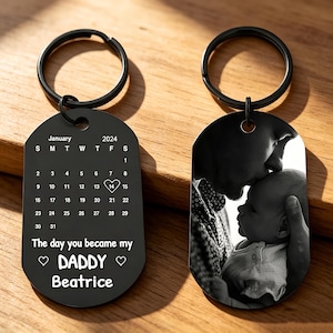 Op de afbeelding: Twee zwarte metalen sleutelhangers. Eén sleutelhanger heeft een kalender voor januari 2024 met een hartje op de 14e en de tekst "The day you became my DADDY Beatrice". De andere heeft een zwart-wit foto van een ouder die een baby kust.
