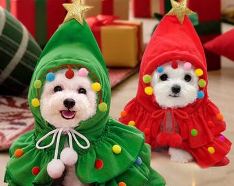 May include: Two small white dogs wearing Christmas tree costumes. One dog is in a green costume with a gold star on top, colorful pom-poms, and a white tie. The other dog is in a red costume with a gold star and colorful pom-poms.