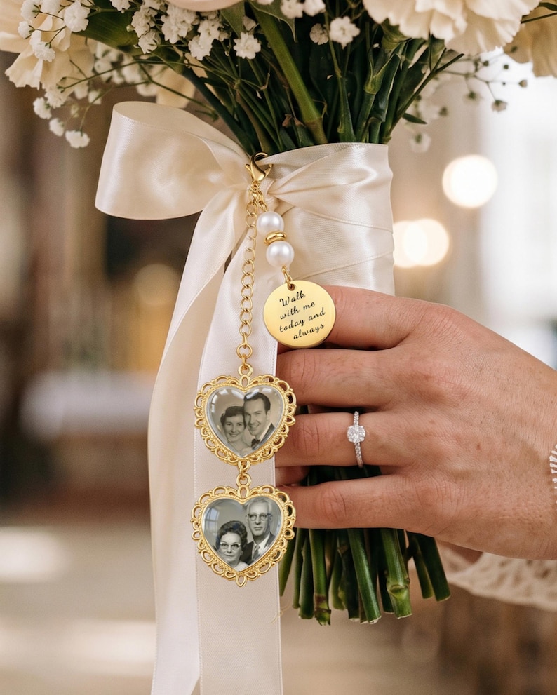 Op de afbeelding: Een goudkleurige metalen bedel bevestigd aan een cr&egrave;mekleurig satijnen lint op een bloemenboeket. De bedel bevat twee hartvormige lijstjes met zwart-witfoto's en een ronde schijf met de inscriptie Walk with me today and always.
