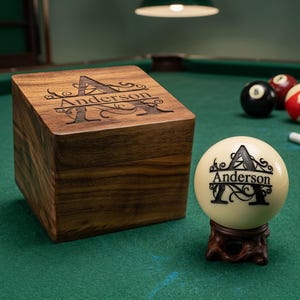May include: A wooden box and a billiard ball with the name "Anderson" engraved on them. The box is square and has a lid with the same engraving. The billiard ball is cream-colored and rests on a small wooden stand. Other billiard balls are in the background.