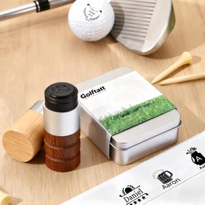 May include: A golf-themed gift set featuring a wooden and metal golf ball marker, a metal box with the text "Golftatt", golf tees, a golf ball, and a golf club. The golf ball marker has a black top and a wooden base.