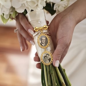 Op de afbeelding: Een goudkleurige metalen broche met twee ovale frames met ingewikkelde filigraanranden, elk met een zwart-witfoto. De broche is bevestigd aan een wit satijnen lint dat om de stelen van een boeket witte bloemen is gewikkeld.