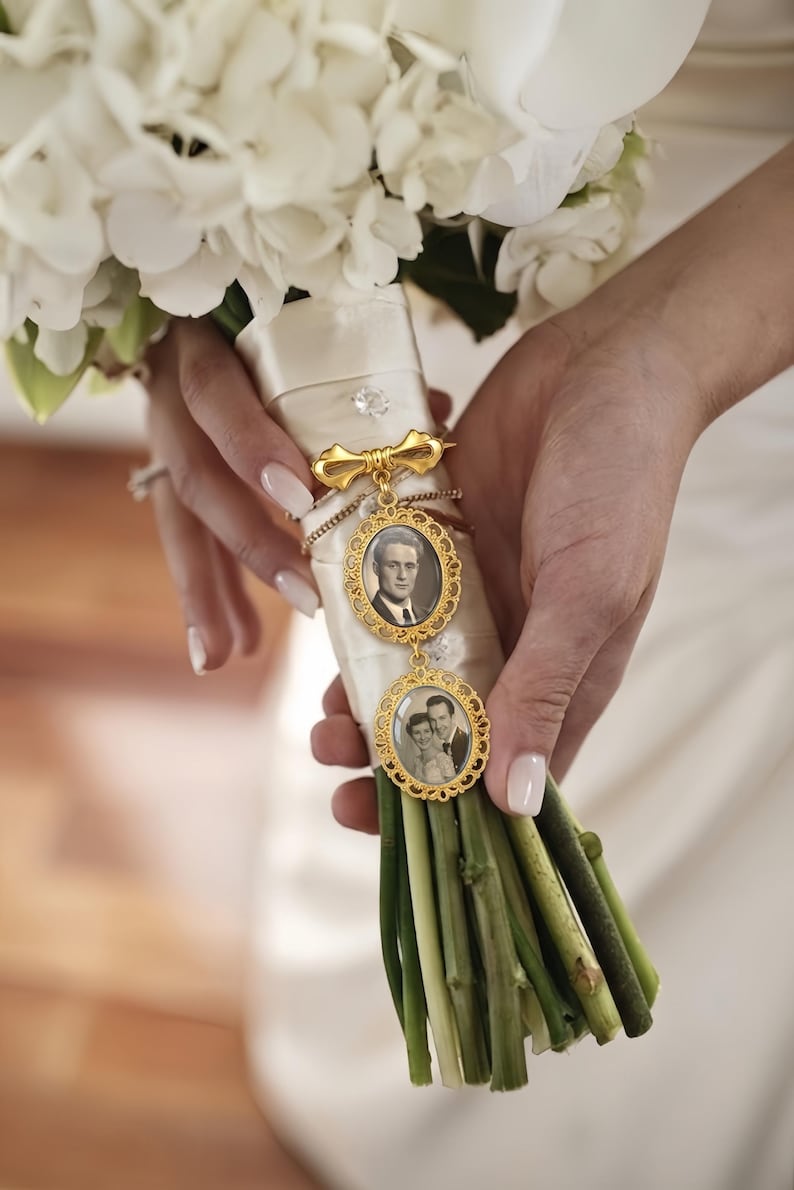 Op de afbeelding: Een goudkleurige metalen broche met twee ovale frames met ingewikkelde filigraanranden, elk met een zwart-witfoto. De broche is bevestigd aan een wit satijnen lint dat om de stelen van een boeket witte bloemen is gewikkeld.