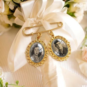 May include: A gold-toned safety pin brooch featuring two oval-shaped pendants with ornate filigree frames. Each pendant contains a black and white portrait photograph of a person, secured to a white satin ribbon wrapped around a floral bouquet.