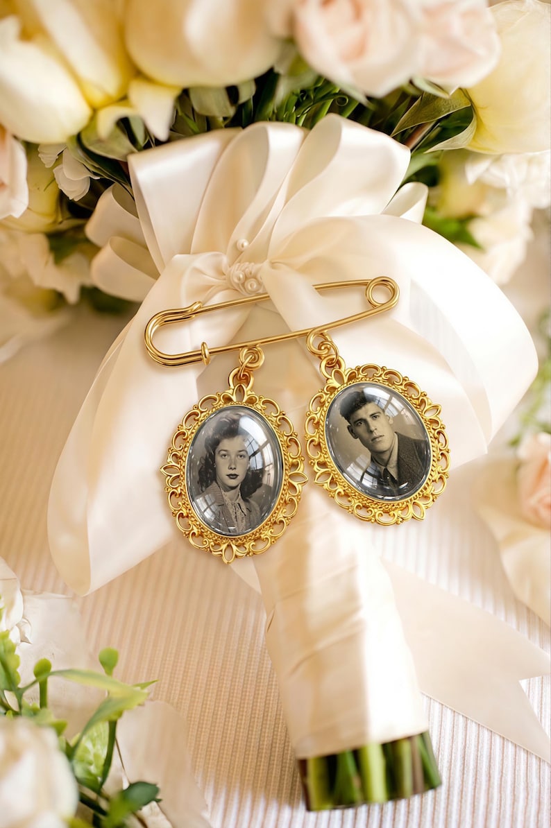 May include: A gold-toned safety pin brooch featuring two oval-shaped pendants with ornate filigree frames. Each pendant contains a black and white portrait photograph of a person, secured to a white satin ribbon wrapped around a floral bouquet.