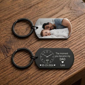 May include: Two black keychains with rectangular pendants. One pendant displays a photo of a man and a baby. The other pendant is black with a clock design, the text "The moment you became my DAD", date, time, and a name.