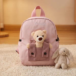 May include: A light pink backpack featuring the name Victoria embroidered in white thread. The front includes a textured mauve pocket containing a plush beige teddy bear. A separate, detached plush beige rabbit sits beside the backpack.