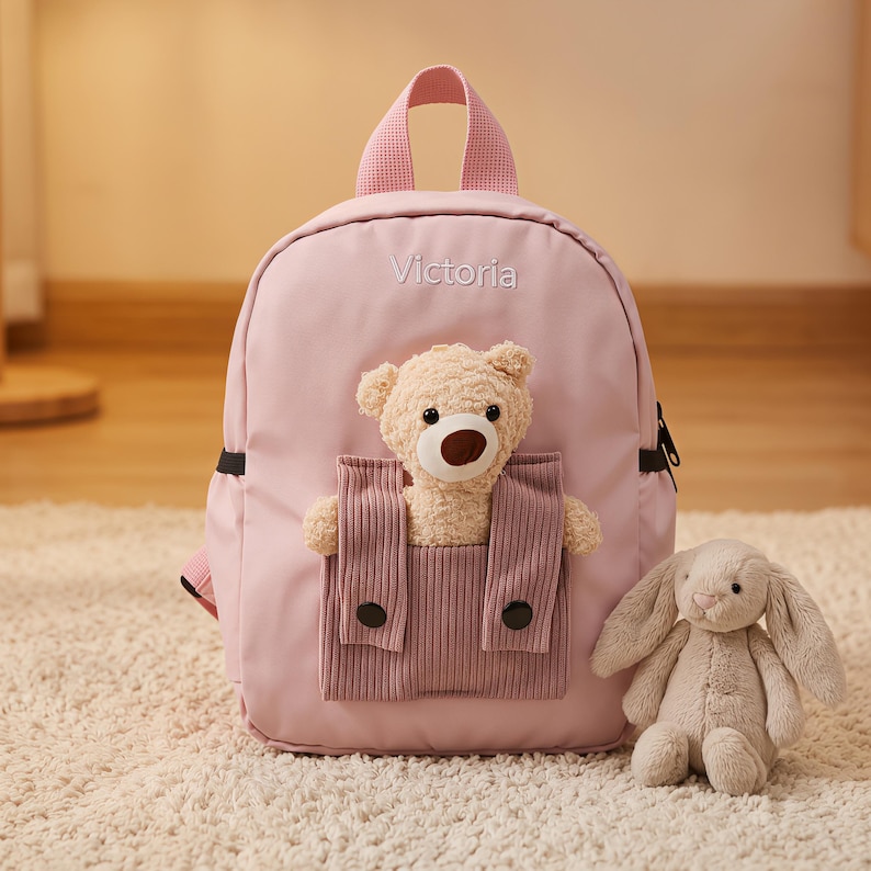 May include: A light pink backpack featuring the name Victoria embroidered in white thread. The front includes a textured mauve pocket containing a plush beige teddy bear. A separate, detached plush beige rabbit sits beside the backpack.