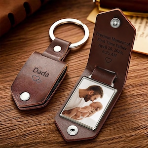 May include: A brown leather keychain with a silver key ring. The keychain has the word "Dada" and two hearts engraved on one side. The other side opens to reveal a photo and the text "Precious Moment of First Time Father April 20, 2026, 6:25 AM."