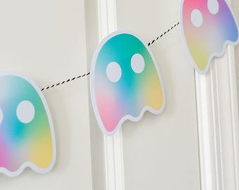 Puede incluir: Una colorida guirnalda con forma de fantasma con un degradado de color turquesa, rosa y amarillo. Cada fantasma tiene dos ojos circulares blancos y está ensartado en un cordel blanco y negro. La guirnalda es una decoración festiva.