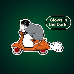 May include: A sticker featuring a cartoon opossum riding an orange scooter. The opossum wears a black helmet. The sticker has a white border and the text "Glows in the Dark!" in a white oval. The scooter has the word "motohop" on it.