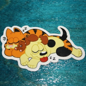 May include: A cartoon sticker featuring a sleeping orange tabby cat with black stripes cuddling a sleeping yellow puppy with black spots and stripes. The cat has a pink nose and the puppy has a red tongue. Both animals have closed eyes and are making "zz" sounds.
