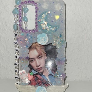 Peut inclure: Une coque de t&eacute;l&eacute;phone transparente avec un motif bleu clair et blanc. La coque pr&eacute;sente une photo d'un homme aux cheveux blonds, portant une chemise &agrave; carreaux bleus et rouges et une veste en jean bleue. La coque comprend &eacute;galement un petit lapin blanc avec une carotte rouge, une fleur bleue et une paire d'ailes blanches. La coque est dot&eacute;e d'une sangle perl&eacute;e avec le mot "MOONBEBE" dessus.