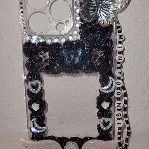 Peut inclure: Une coque de t&eacute;l&eacute;phone transparente avec un motif noir et blanc. La coque est orn&eacute;e d'un charme papillon, d'une cha&icirc;ne de perles et de perles noires, et d'une paire d'ailes blanches. La coque a &eacute;galement un dos transparent avec une finition scintillante.
